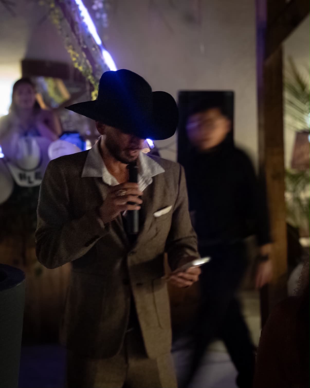 Cowboy-hatted host speaking into a microphone among Old Money guests